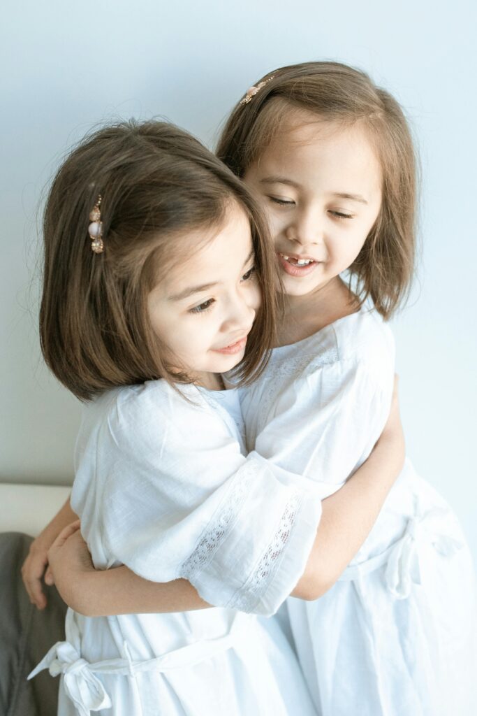 Cute moment of two young girls embracing indoors in white dresses.